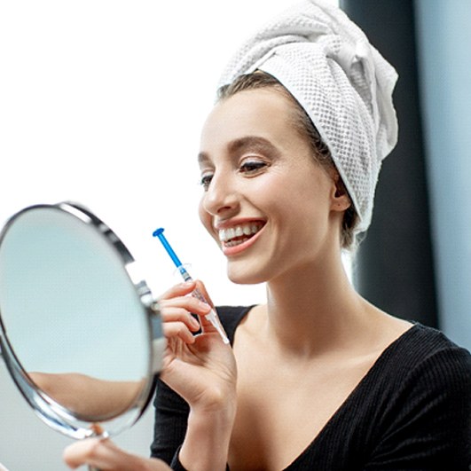 A young woman with a towel on her head, holding a syringe with whitening gel and a hand-held mirror to look at her brighter smile